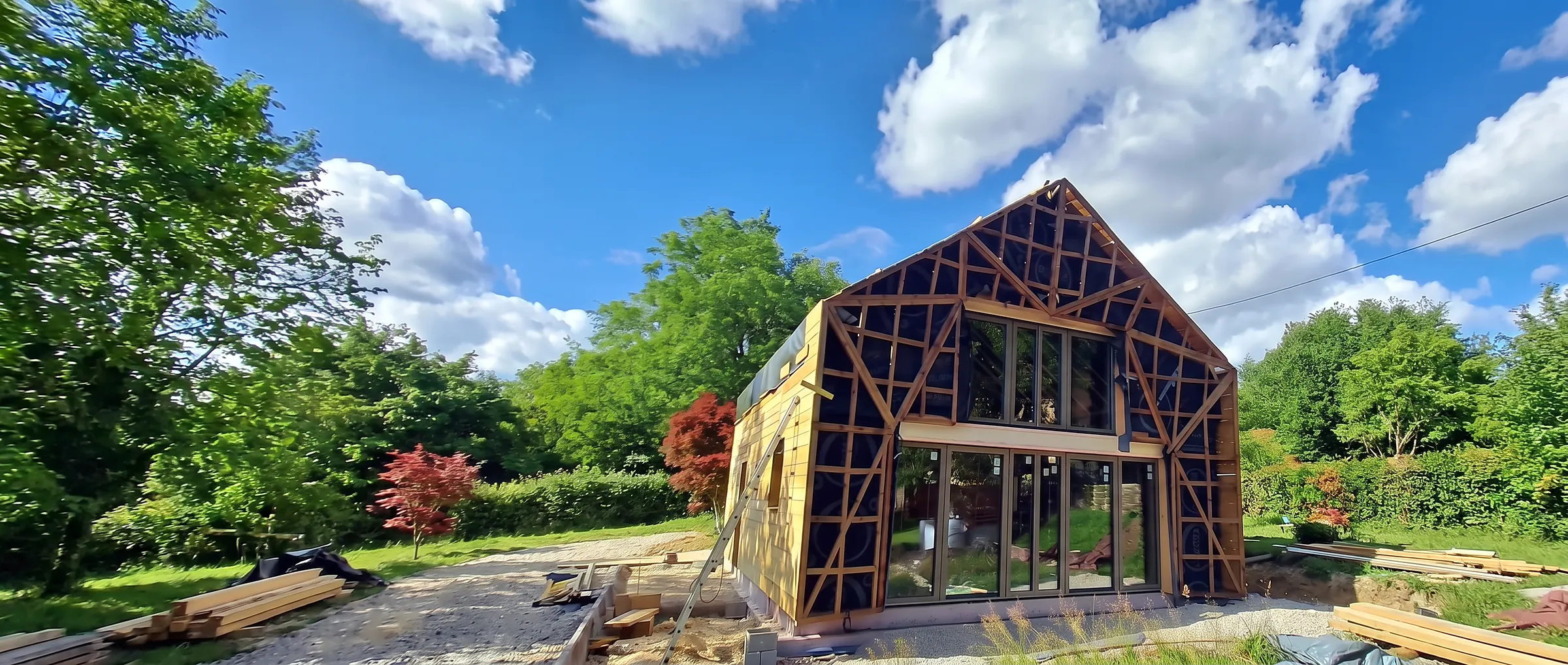 Maison à ossature bois en cours de construction, charpente et bardage bois apparents, sous un ciel bleu dans le Jura